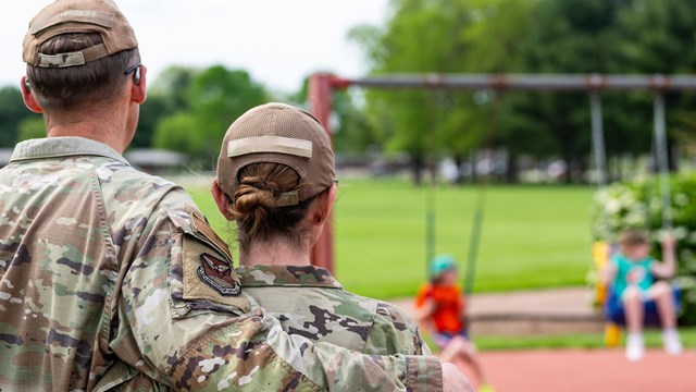 While watching two kids swing, a man and woman in uniform wrap their arms around one-another.