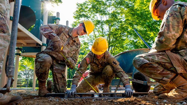 3 men in hard hats and military uniforms use a level and measuring tape at the foot of a playground.