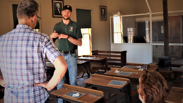 A tall young man speaks with people standing in a room filled with wooden school desks.