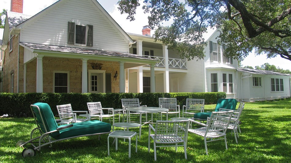 White lawn chairs sit in a circle on the lawn in front of a large, two-story white home.