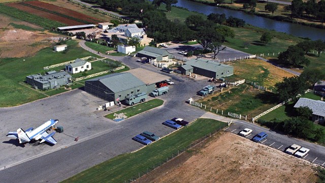 Aerial view of several green structures, white and blue jet, and 1960s cars alongside a river.