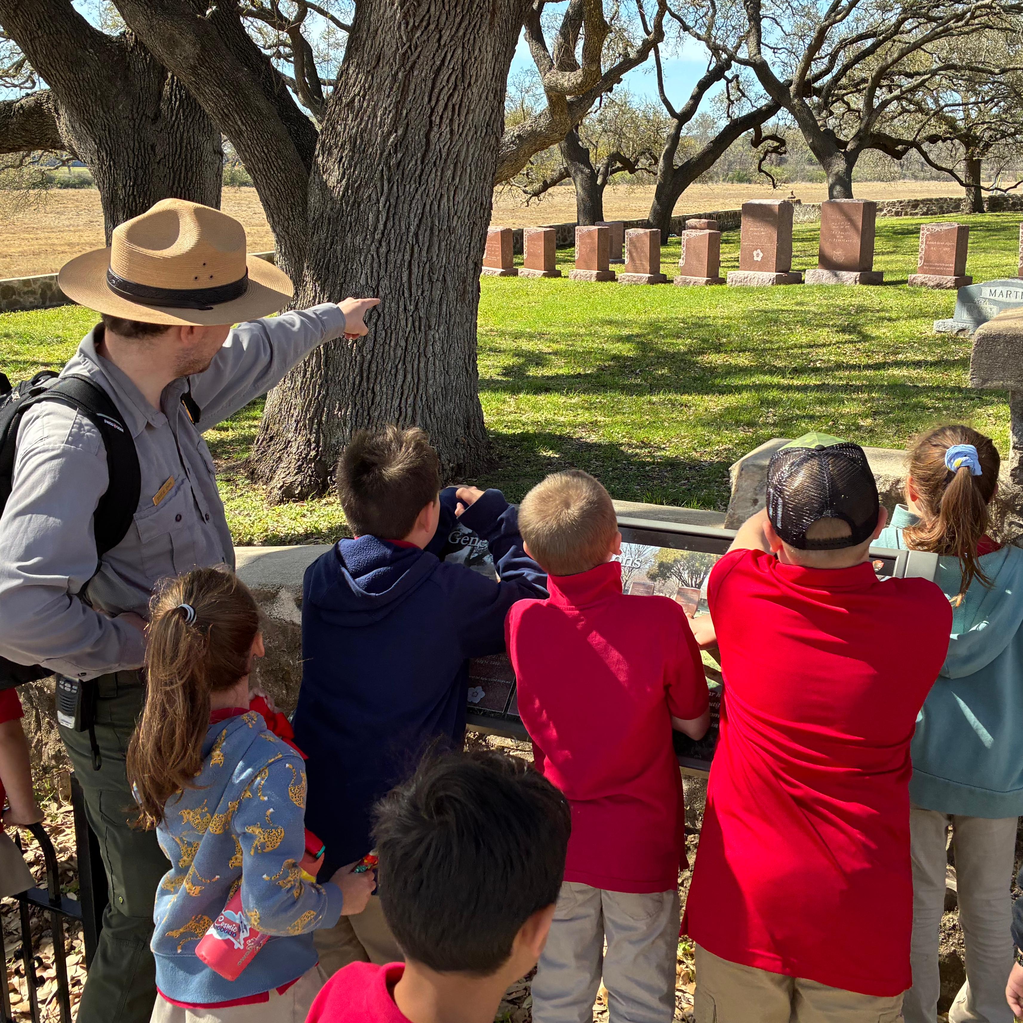 A park ranger points over a cemetery wall while surrounded by children who all look on. 