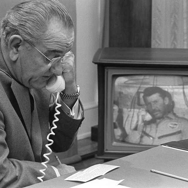President Johnson speaks with a man on a phone and television. 