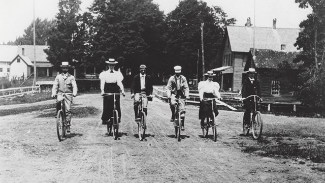 historic black and white photo of group of 6 people on bicyles on dirt road