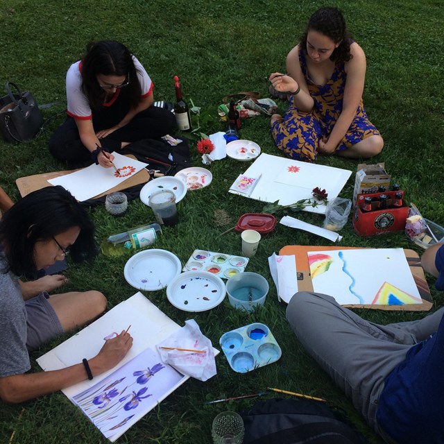 group of people sit on a grassy lawn painting watercolor