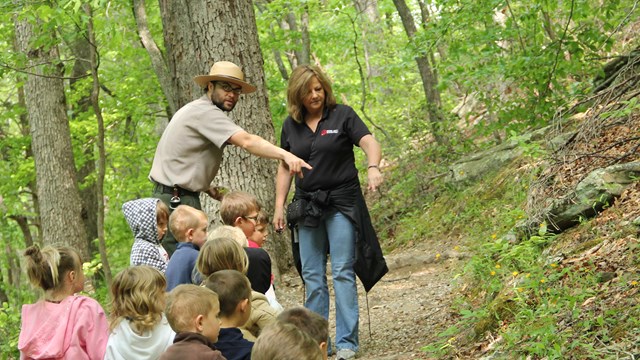 A park ranger points out wildflowers to a visiting school group while hiking along a forest trail.