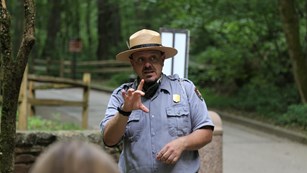 A park ranger answers visitors' questions while outdoors, in a forest setting.