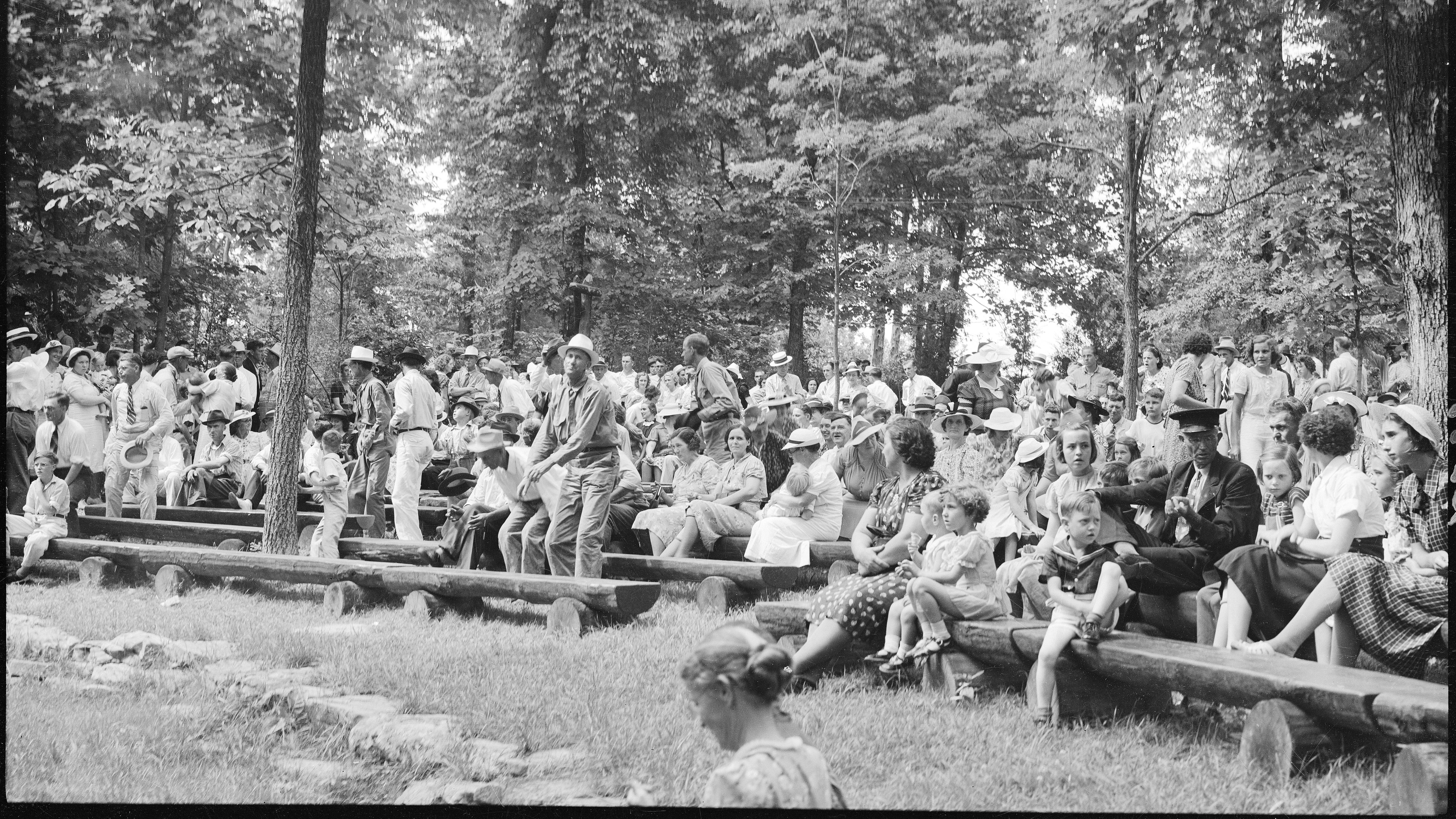 A large group of people seated on wooden benches in an outdoor setting.