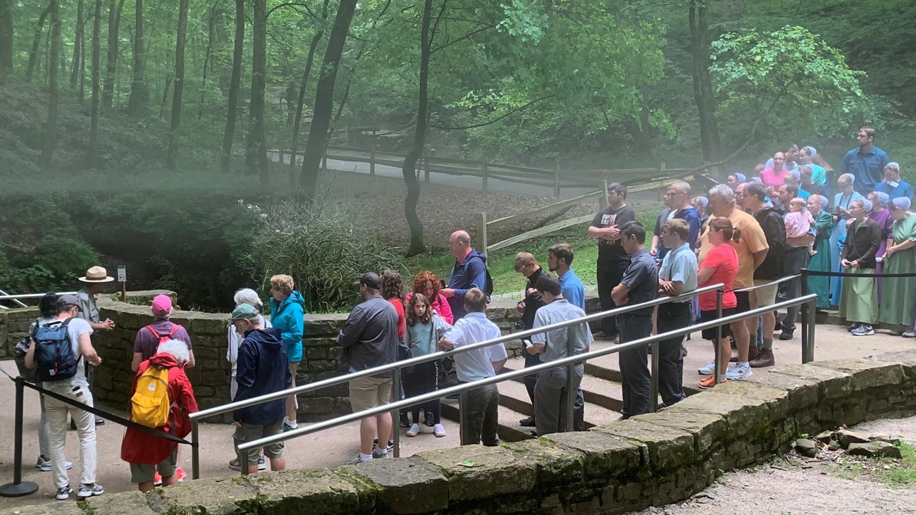 A large group of people stand at the entrance to a cave in a forest.