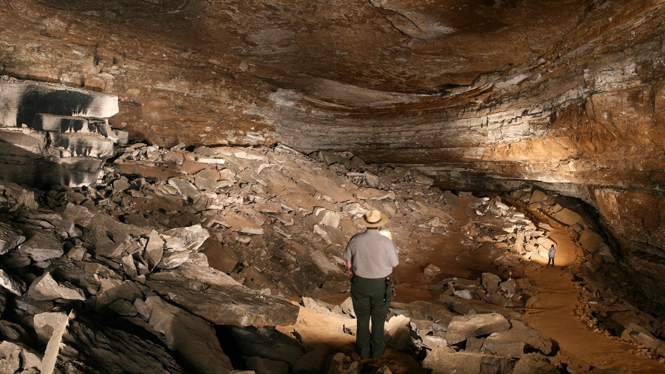 A park ranger standing in the middle of a large cave room with broken rocks all along the floor. 