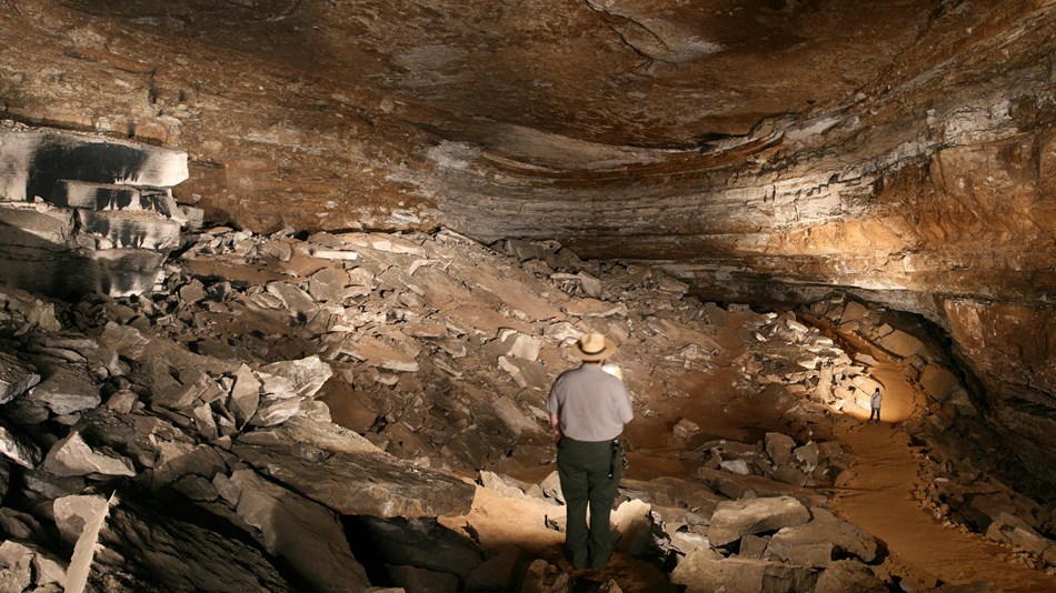 A park ranger standing in the middle of a large cave room with broken rocks all along the floor. 