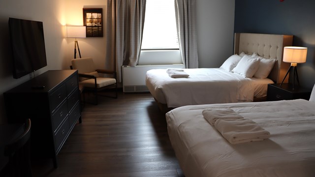 A hotel room with two beds softly illuminated by a window and lamp.