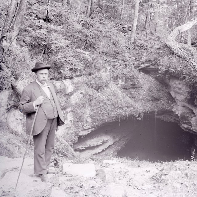 A black and white photo of a man standing at the cave entrance 