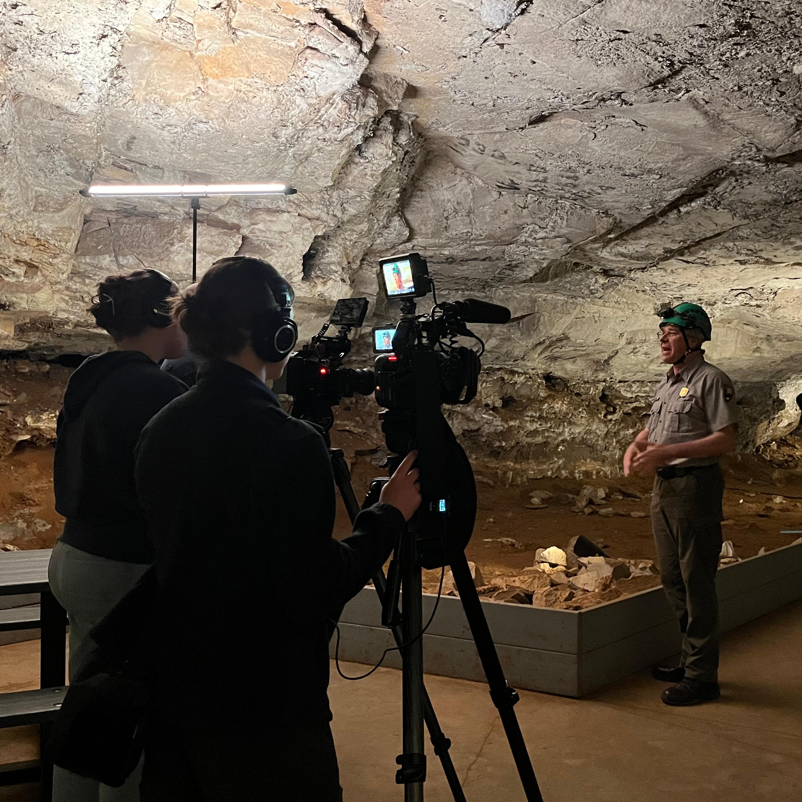 A park ranger wearing a helmet speaks to a film crew with cameras and lights in a large cave.