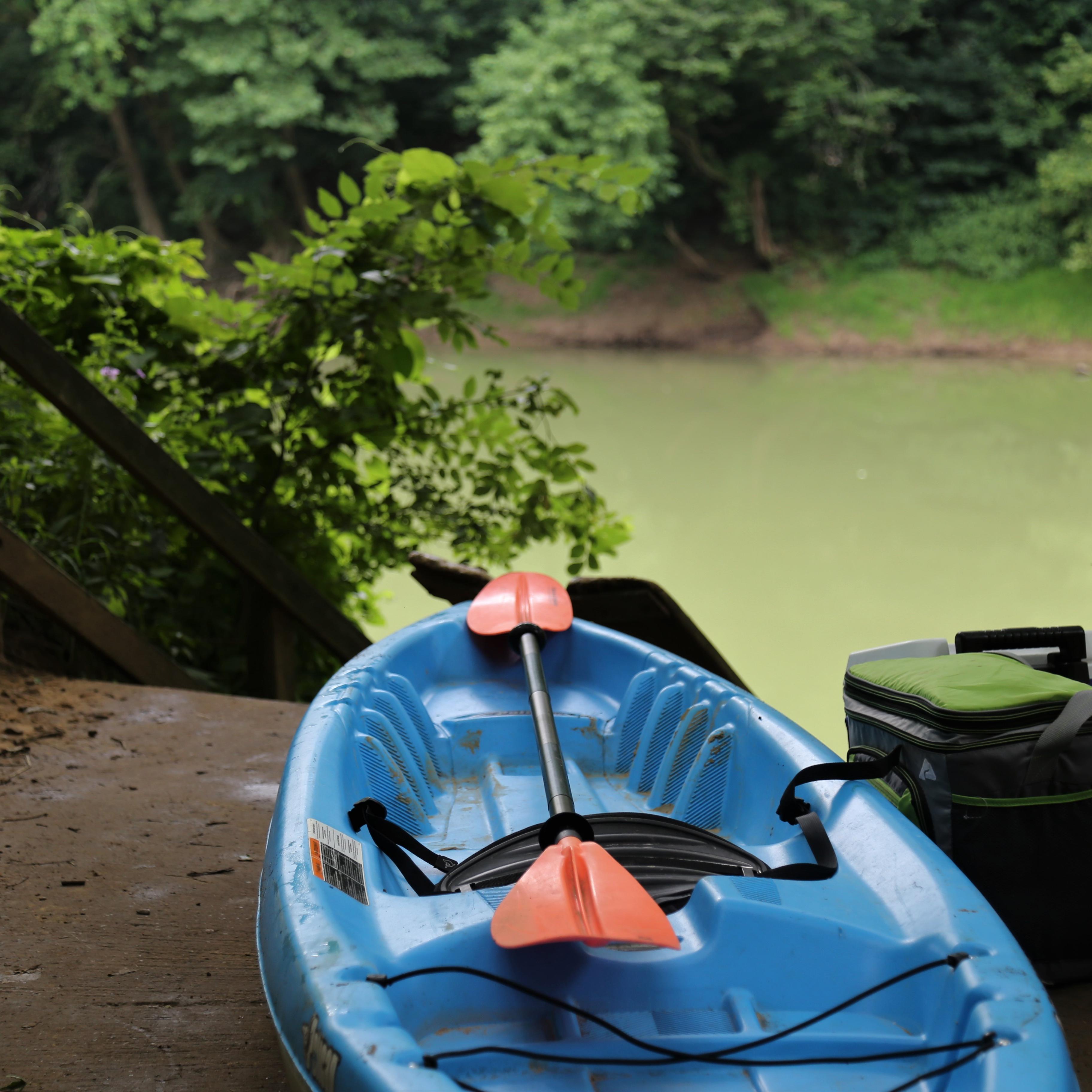 A blue kayak with a paddle rests at the top of a ramp that descends into a wide green river.