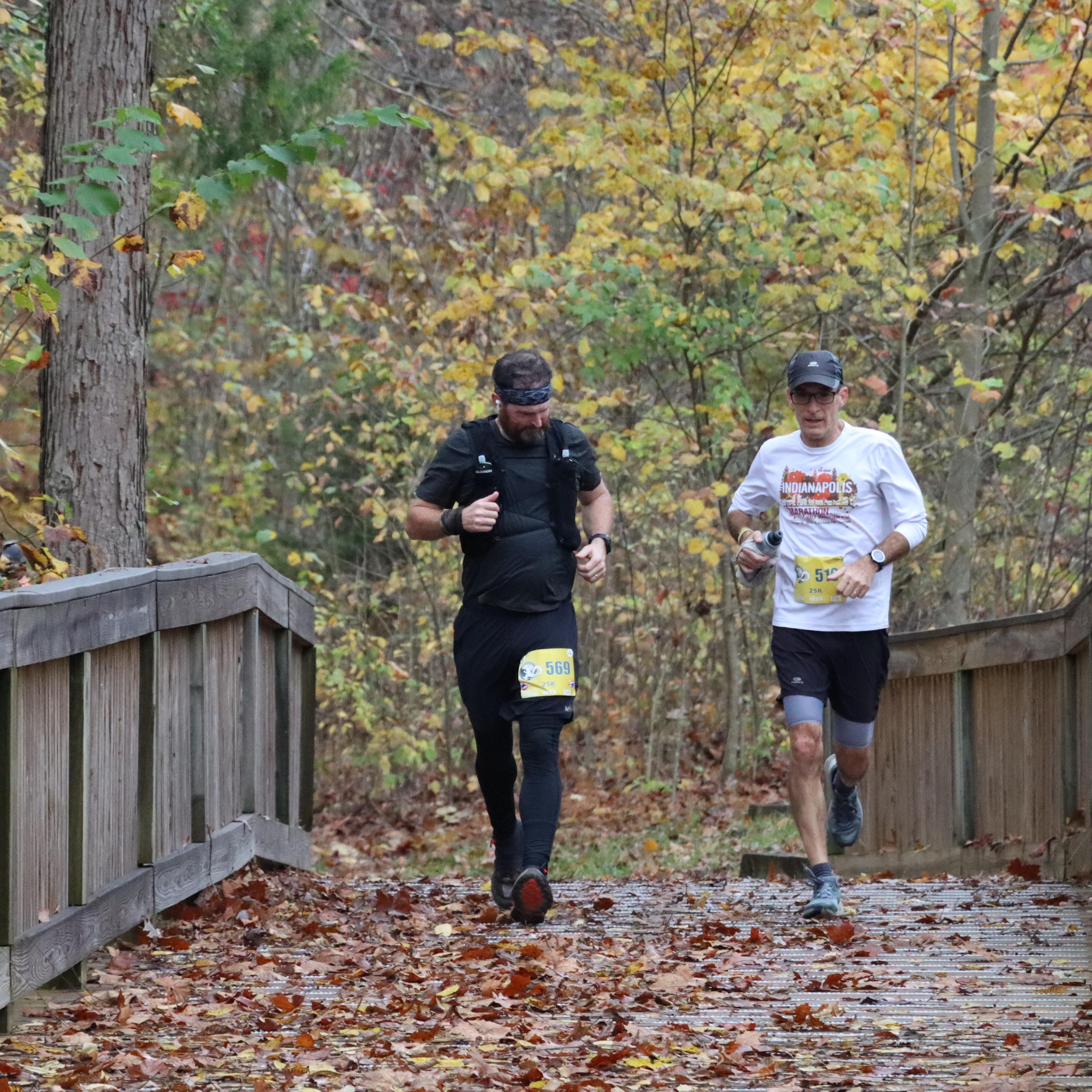 Two people wearing race bibs with numbers run on a leaf covered trail.