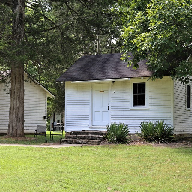 Small wood cabins with white paint. Tall trees and green grass surrounds them. 