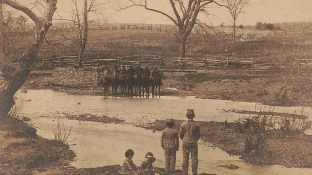 Sepia tone photo shows four children in foreground and soldiers mounted on horseback across creek