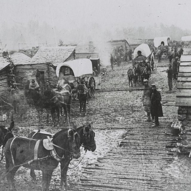 Black and white photo of people and horses standing around a camp with wooden cabins and tents.