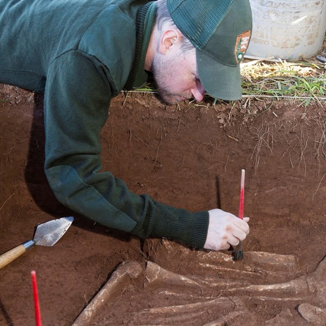 Color photo of an NPS employee laying on the ground brushing dirt away from an archeology excavation