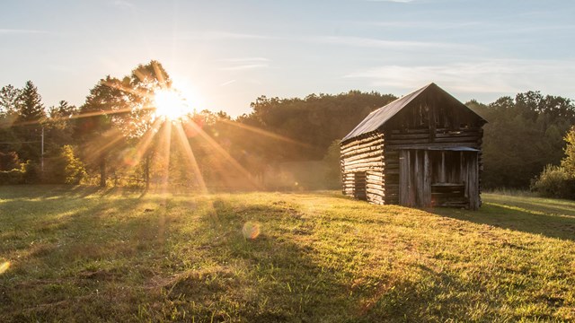 A cabin in a field with sunlight behind it.