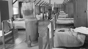 A black and white photo of a bunk house with beds lining the sides of the room.