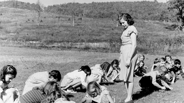 A woman stands in a field with 16 kids crouching in the dirt around her.