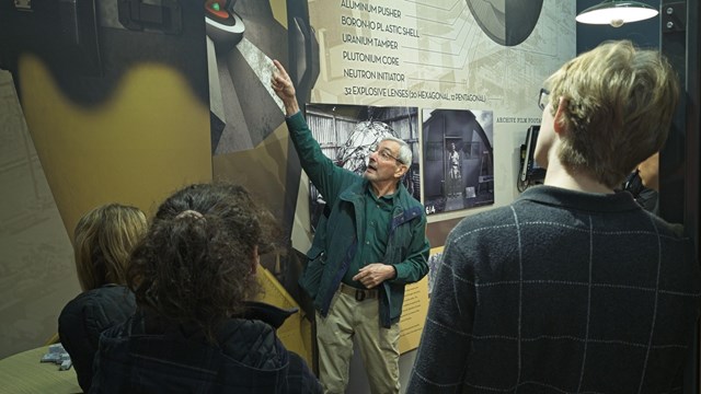 A man points at a display of a bomb core on the wall while 3 students watch.