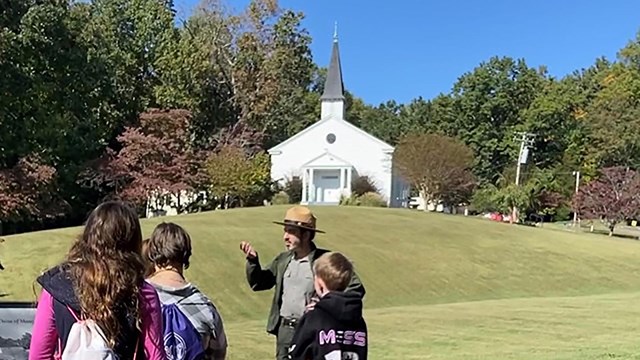 A uniformed ranger talks with 3 people outside in the sun in front of a white church.