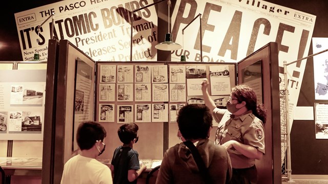 A black and white photo of kids talking to a speaker who is pointing out newspaper clippings.