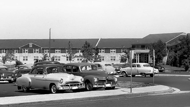 A black and white image of cars in the parking lot of a building.