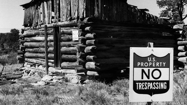 A black and white photo of a rustic, log cabin with a black and white sign that reads US government.