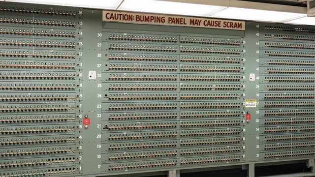 A wall of knobs with text at the top that reads “Caution: Bumping Panel May Cause Scram.”