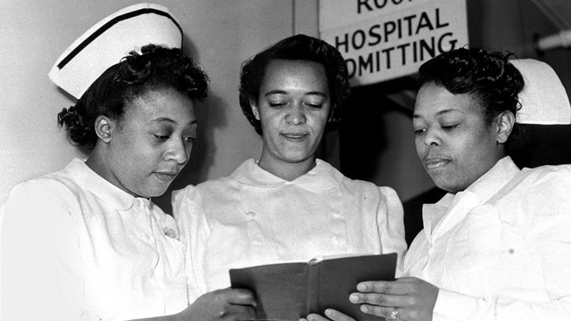 Black and white photo of three women in nurses uniforms looking at a book. 