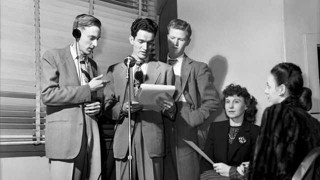 Black and white photo of three men and two women gathered around a microphone.