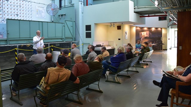 Several people seated on benches watching a speaker in front of a reactor face. 