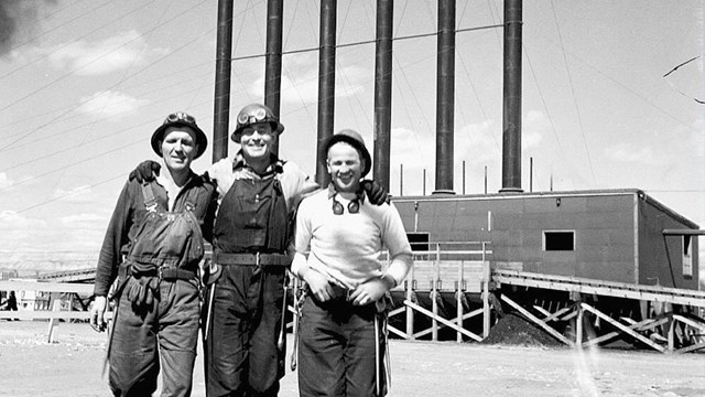 A black and white photo of three workers in front of smoke stacks.