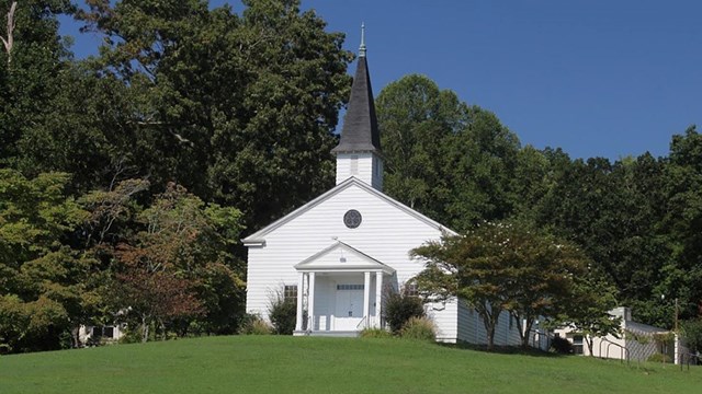 Small white chapel with a tall steeple on a grassy hill with trees behind it on a sunny day 