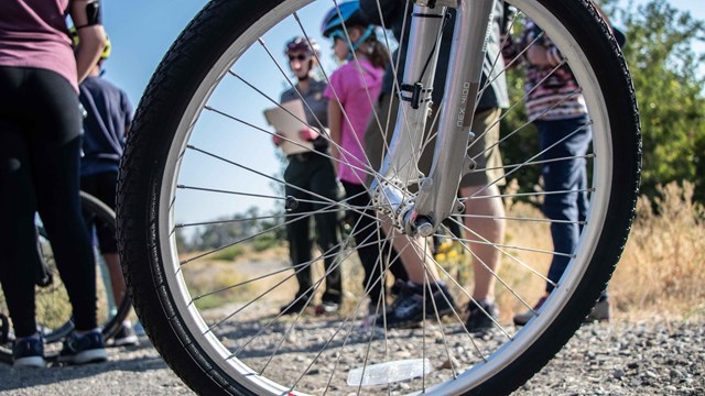 A group of people with bike helmets can be seen through the spokes of a bicycle wheel.