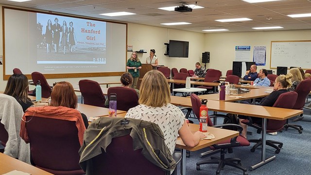 10 adults sit at tables looking at 2 women in front of a screen that reads "The Hanford Girl."