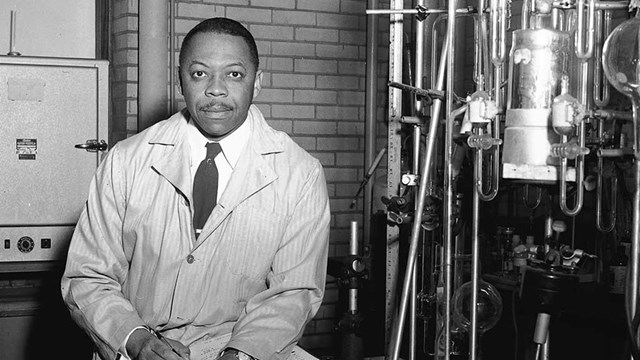 A black and white photo of a man in a white coat next to equipment.  