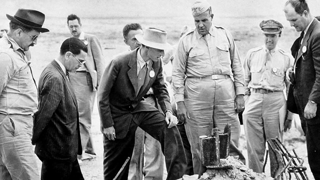 A black and white photo of men in suits and uniforms looking at something on the ground. 