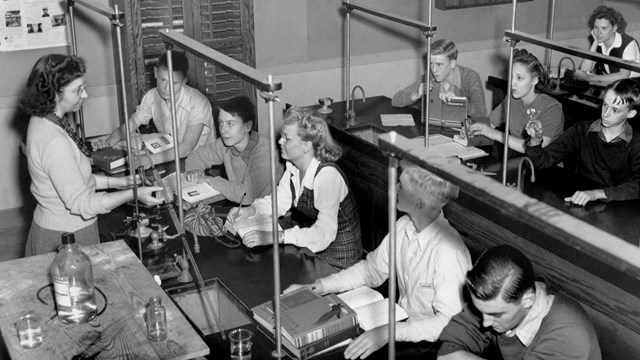 Black and white photo of 9 students sitting at desks listening to a teacher.