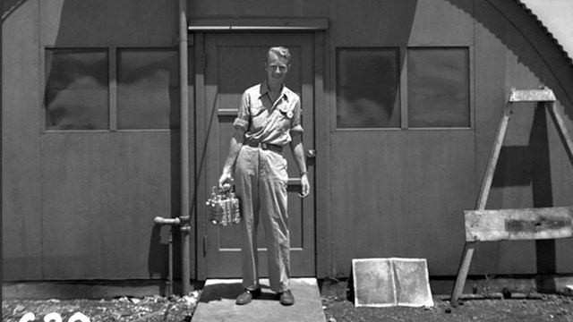 A black and white image of a man standing in front of a rounded hut.