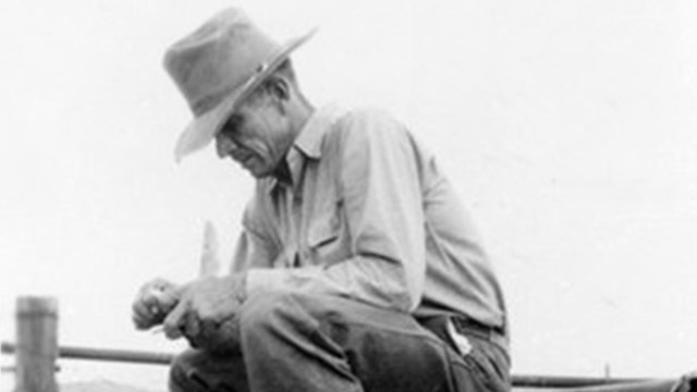 A black and white image of a man in a light-colored shirt and dark pants with a wide-brimmed hat sit
