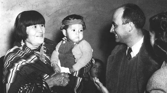 A black and white image of a lady with short black hair holding a baby in front of a couple. 