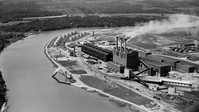 Black and white photo of a several industrial-looking buildings along a river. 