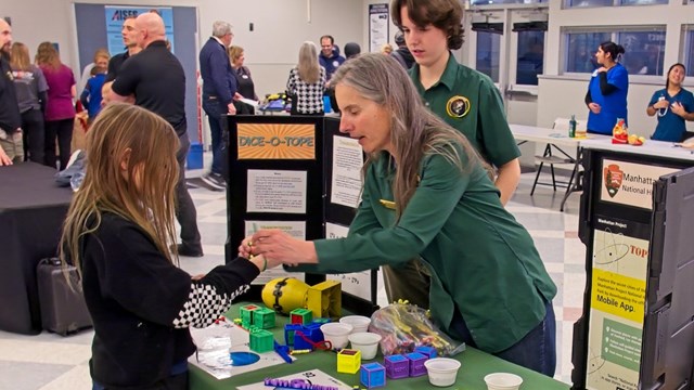 A woman leans over a table ties a bracelet on a young girl’s wrist who is facing the table.