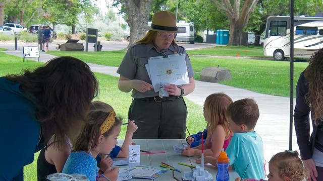 A uniformed woman stands at the end of a table holding paper. 7 kids work on books at the table. 