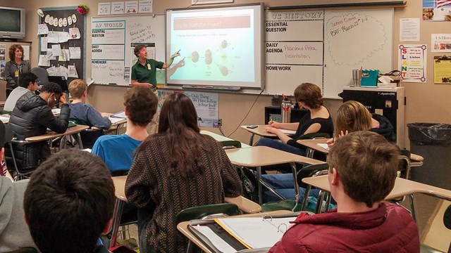 9 students sit at desks listening to a woman pointing at a screen displaying fission. 